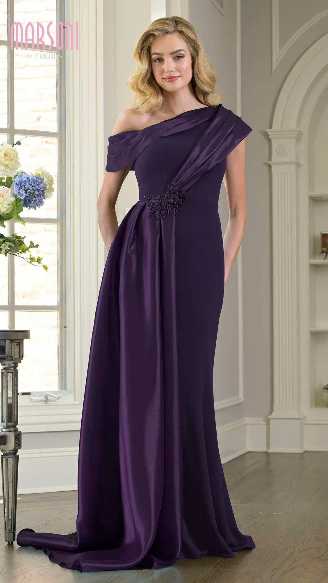 A woman stands indoors wearing the Marsoni M346 Long Formal Dress in eggplant, an off-the-shoulder, floor-length stretch crepe gown featuring floral waist detailing and an elegant sheath silhouette.